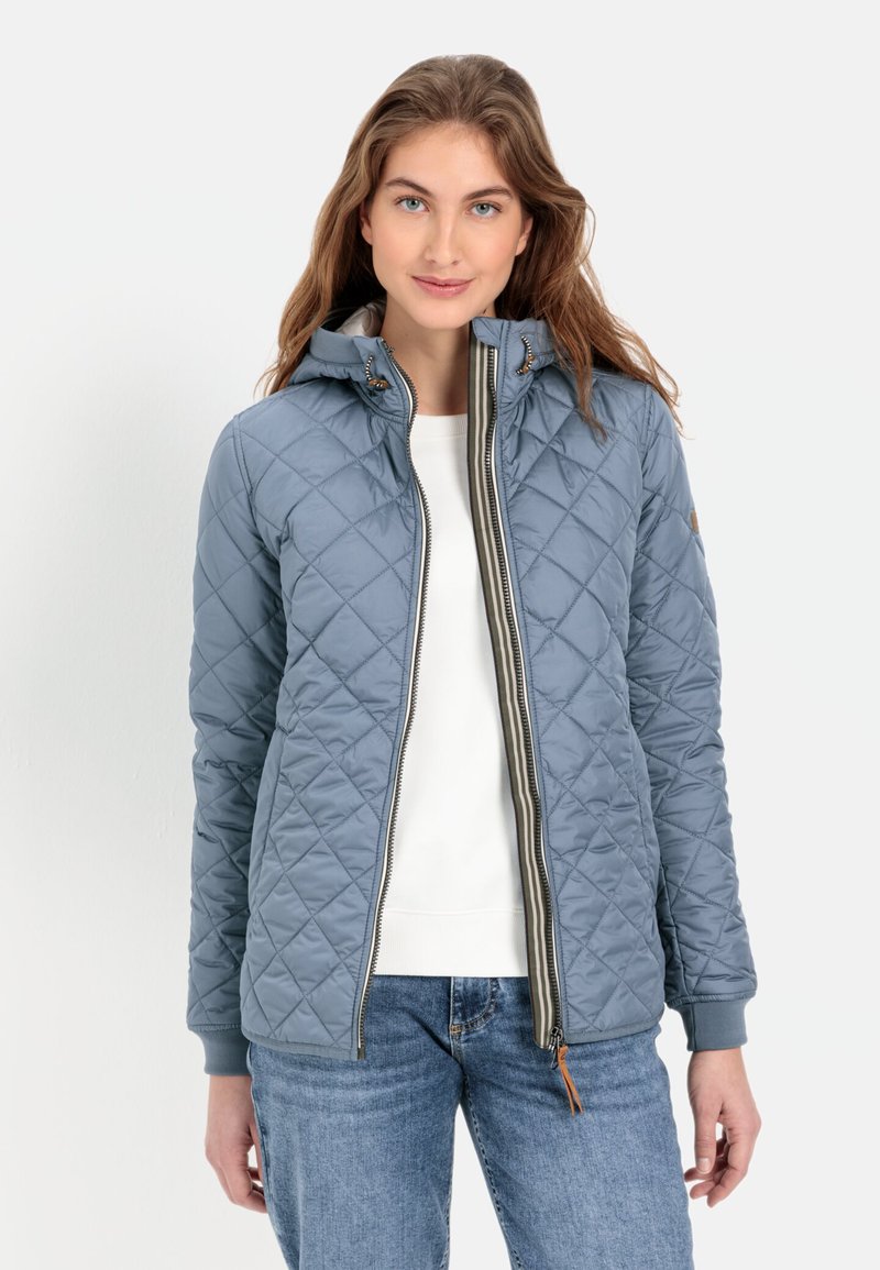Quilted blauwe jas met capuchon, voorzien van een ritssluiting aan de voorkant, een gegerande textuur en ribgebreide manchetten. Gepaard met een witte top en jeans.