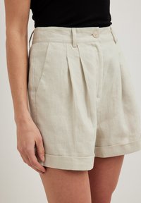 Plisserade shorts i ljusbeige linne, med en midja med knappstängning, fickor på sidan och uppvikta fållar. Slät textur.