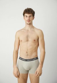 Graue Baumwoll-Boxershorts mit schwarzem elastischen Bund, der mit dem "JACK & JONES"-Logo versehen ist. Das Modell hat kurze lockige Haare und steht vor einem hellen Hintergrund.
