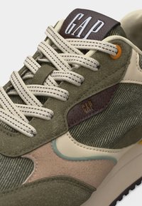 Αθλητικό παπούτσι σε ελιά πράσινο και μπεζ με λευκά κορδόνια, υφή υφάσματος και λογότυπο "GAP" στη γλώσσα και στο πλευρικό πάνελ.