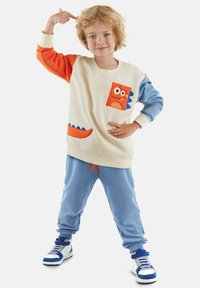 Sweatshirt met een crèmekleurig lichaam, oranje en blauwe mouwen, en een monster zakontwerp. Gecombineerd met blauwe joggingbroeken met oranje accenten.