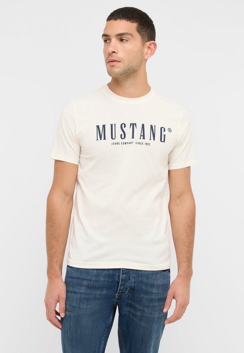 Kremowa koszulka z bawełny, z ciemnoniebieskim logo i napisem "MUSTANG", okrągły dekolt, krótkie rękawy, w połączeniu z niebieskimi dżinsami.