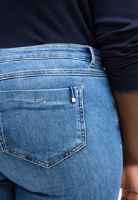 Άτομο που φοράει ανοικτό μπλε τζιν παντελόνι και σκούρο μπλε πουκάμισο με μακριά μανίκια, δείχνοντας την πίσω τσέπη και μέρος του χεριού.