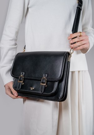 Sac en cuir noir en bandoulière avec fermetures à boucles doubles, accents en métal doré et coutures blanches contrastantes. Texture lisse.