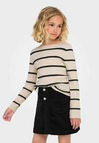 Młoda dziewczyna z kręconymi blond włosami, nosząca beżowy, prążkowany top z długim rękawem w czarne paski oraz czarną spódnicę z korduroyu zapinaną na guziki.