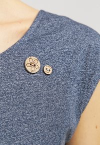 To træknapper i træ, den ene stor med et blomsterdesign og den anden lille med tekst, på et blåt struktureret stof nær en persons skulder.