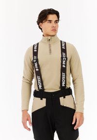 Beige fleecetop met rits en hoge hals, gecombineerd met zwarte broek met beige accenten en merkbare bretels met de tekst "PROTEST".