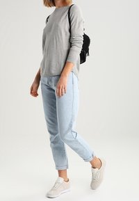 Pull gris clair à manches longues, jean bleu clair retroussé et baskets beige. Un petit sac à dos noir texturé est porté sur une épaule.