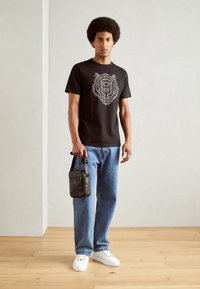 Camiseta de algodón negra con un gráfico de león blanco, acompañada de jeans de denim azul claro y zapatillas blancas. Lleva una pequeña bolsa negra.