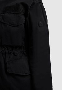 Czarny rękaw kurtki z dwoma kieszeniami z patkami oraz okrągłą naszywką z liściem klonu na górnej części ramienia.