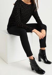 Schwarzes Langarmoberteil mit floralem Muster, kombiniert mit schwarzen Slim-Fit-Hosen und glänzenden schwarzen Stiefeletten mit grobem Absatz.