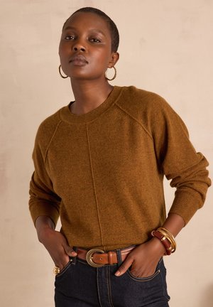 Brauner gestrickter Pullover mit Raglanärmeln, gerippten Bündchen und rundem Ausschnitt, kombiniert mit einem braunen Gürtel und gestapelten Armbändern vor neutralem Hintergrund.