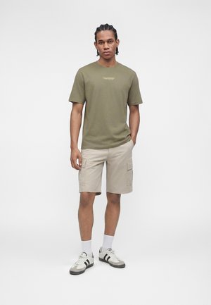 Mężczyzna stojący, ubrany w oliwkowo zielony t-shirt z napisem "California", beżowe krótkie spodnie typu cargo, białe skarpetki i białe trampki z czarnymi paskami.