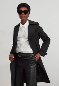 Schwarzer Trenchcoat über einem weißen Hemd, kombiniert mit schwarzen Lederhosen. Große schwarze Sonnenbrillen setzen ein auffälliges Detail. Glatte Texturen überall.