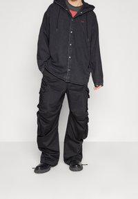 Svart denimskjorta med knappar och huva, bärs över en röd T-shirt. Luftiga svarta cargobyxor med flera fickor. Svarta stövlar.