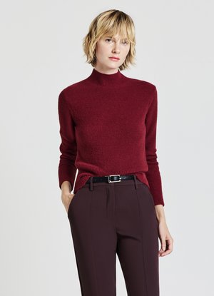 Maglione a coste color bordeaux con colletto alto e polsini a costine, abbinato a pantaloni scuri su misura e una cintura nera con fibbia in argento.
