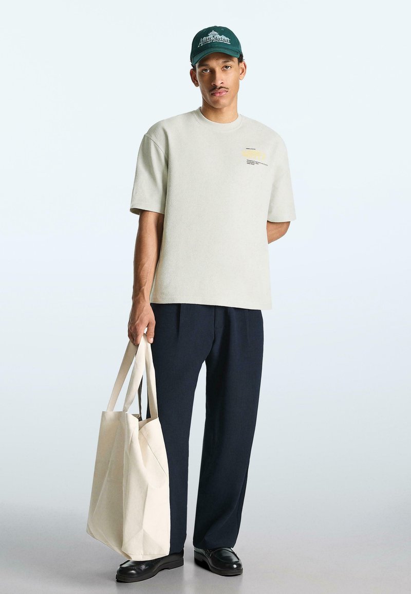 Jeune homme portant un T-shirt oversize beige clair, un pantalon large noir, une casquette vert foncé, des chaussures noires, tenant un grand sac fourre-tout beige.