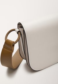 Hvid skuldertaske i læder med glat tekstur, med et rundt accenthåndtag og en brun rem, syninger langs kanterne og et elegant design.