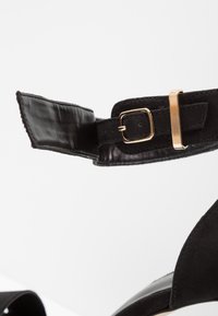 Bride de cheville en daim noir avec boucle et fermoir dorés sur chaussure à talons hauts, montrant la doublure intérieure en cuir et les coutures détaillées.
