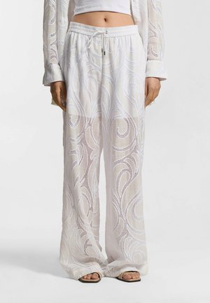 Pantaloni bianchi a gamba larga con intricati motivi di pizzo a spirale, indossati con sandali bassi e un top bianco corto.