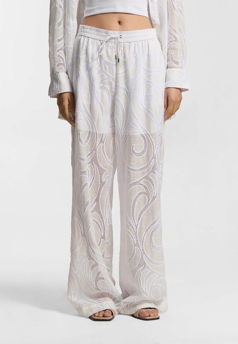 Pantalon blanc à jambes larges avec des motifs complexes en dentelle en forme de tourbillon, porté avec des sandales plates et un haut blanc court.