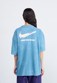 Mėlyna per didelė marškinė pagaminta iš minkštos medžiagos, su dideliu, besišviečiančiu Nike swoosh ir užrašu "GARSAS IŠ PADO" ant nugaros.