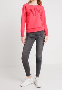 Sudadera de color rosa brillante con logotipo "AX" texturizado, combinada con jeans skinny de color gris oscuro y zapatillas blancas. Tejido suave y diseño informal.