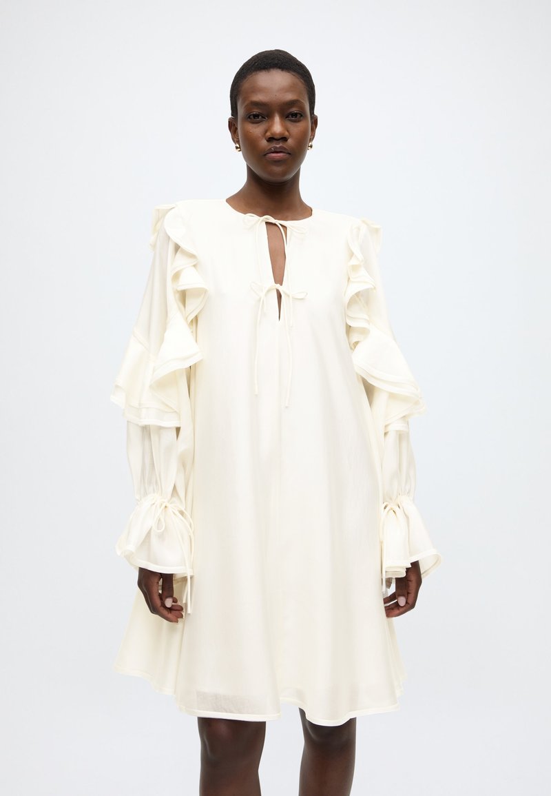 Λευκό φόρεμα με βολάν στα μανίκια και άνοιγμα σε σχήμα κλειδαρότρυπας στο ντεκολτέ. Υφή υφάσματος, μανσέτες που δένονται και άνετη εφαρμογή που δημιουργεί μια ρέουσα σιλουέτα.