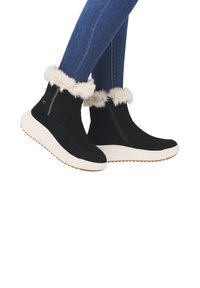 Bottes noires à la cheville avec un revers en fausse fourrure, semelles en caoutchouc blanches et fermeture éclair sur le côté ; associées à un jean skinny bleu.