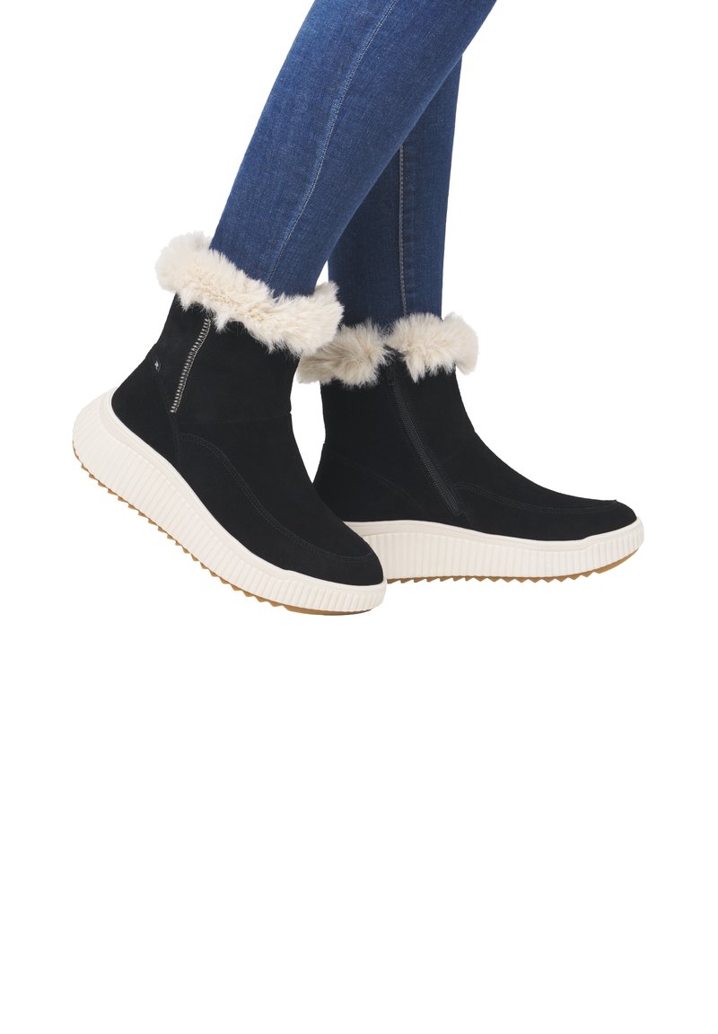 Bottes noires à la cheville avec un revers en fausse fourrure, semelles en caoutchouc blanches et fermeture éclair sur le côté ; associées à un jean skinny bleu.