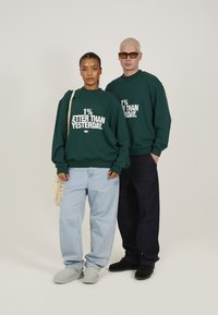 Grønne hettegensere med hvit tekst, blå baggy jeans, og grå og svarte sko. En modell holder en heklet veske. Enkel, avslappet design.