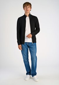 Schwarze, gestrickte Zip-Jacke mit einem strukturierten Muster, getragen über einem weißen T-Shirt, kombiniert mit blauen Jeans und weißen Sneakers.