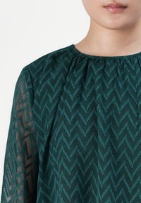 Woman wearing a sheer dark green blouse with textured zigzag pattern and gathered neckline, visible from neck to upper chest.