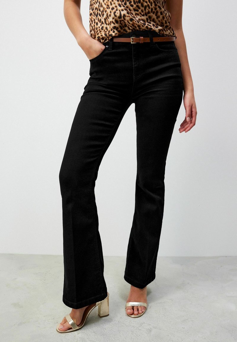 Femme portant un jean flare taille haute noir avec une ceinture marron, un haut imprimé léopard et des sandales à talons blocs dorés à bout ouvert, debout sur un sol gris.