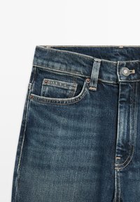 Jeans in denim blu scuro con tasche frontali, passanti per cintura e una chiusura con bottone metallico visibile su uno sfondo bianco.
