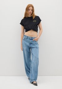 Camiseta negra corta con mangas enrolladas y texto dorado, combinada con jeans de pierna ancha de color azul claro y zapatos negros de punta con acentos de tachuelas.