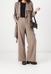 Beżowa, długa, zwiewna kurtka z falbaniastym przodem i otwartym fasonem, w zestawieniu z dopasowanymi spodniami o szerokich nogawkach i brązowymi botkami za kostkę. Ciemna torba.