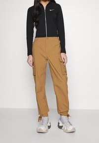 Svart zip-up idrottsjacka, beige cargobyxor med sidofickor, och grå sneakers med vita detaljer. Enkel design med funktionella detaljer.