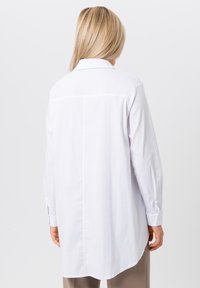 White long-sleeve shirt made of cotton. Features a pointed collar, button cuffs, a back yoke, and a curved hem. Simple and modern design.