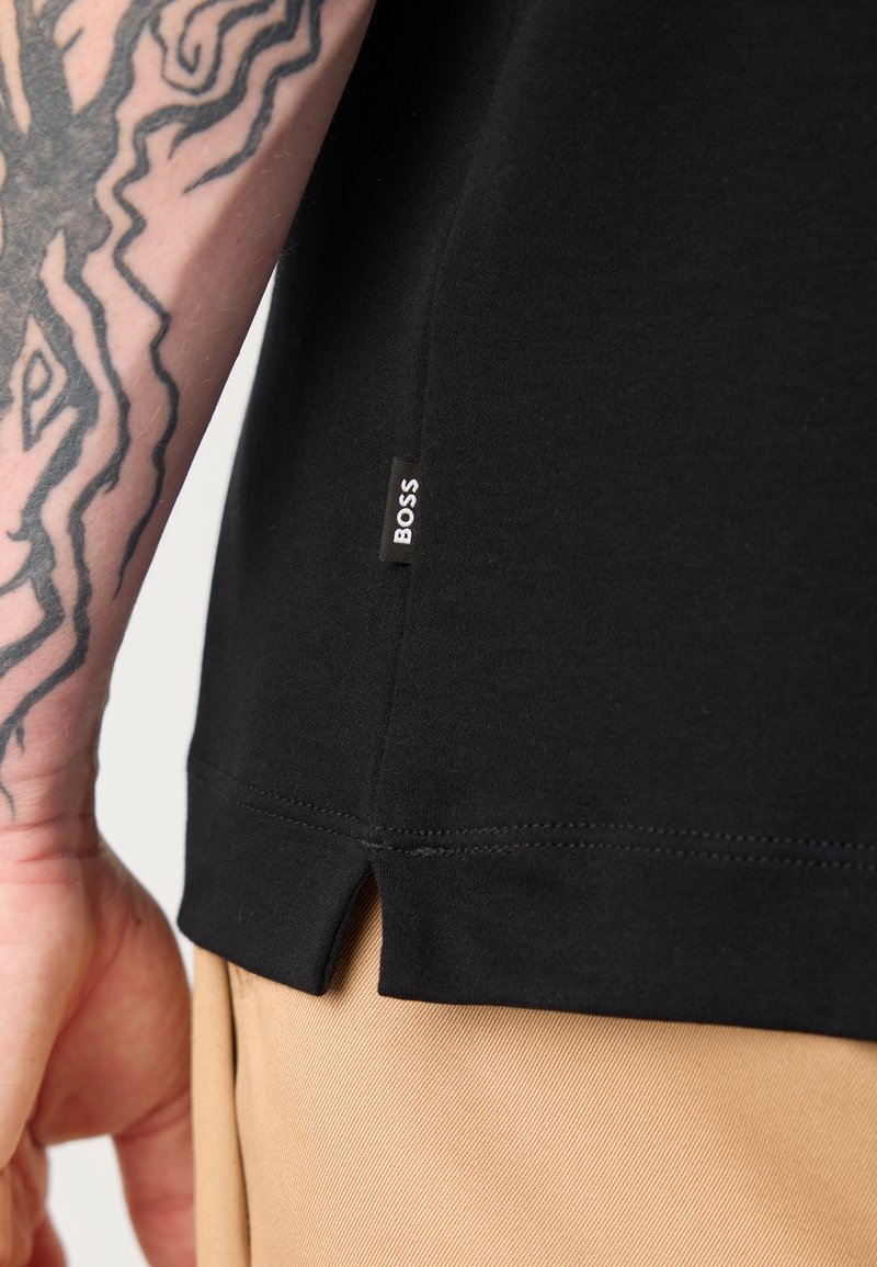 Schwarzes T-Shirt mit seitlichem Schlitz, aus weichem Material. Kleiner Aufnäher mit "BOSS"-Logo am Saum sichtbar. In der Nähe beige Hosen.