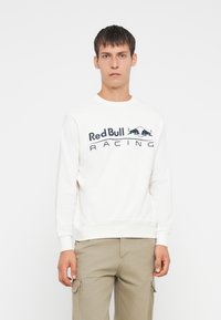 Fiatal ember rövid hajjal, fehér Red Bull Racing kapucnis pulóverben és bézs cargo nadrágban, egy sima fehér háttér előtt áll.