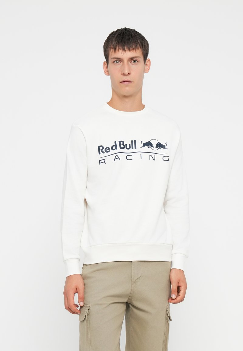 Fiatal ember rövid hajjal, fehér Red Bull Racing kapucnis pulóverben és bézs cargo nadrágban, egy sima fehér háttér előtt áll.