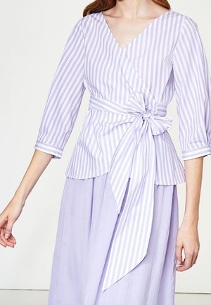 Femme portant un chemisier cache-cœur rayé violet clair à manches trois-quarts noué par un grand nœud à la taille et une jupe assortie.