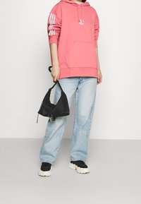 Růžová mikina s kapucí a přední kapsou, má tři bílé pruhy na rukávech a malé logo. Kombinováno se světle modrými džíny a černými teniskami.