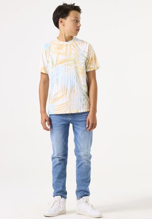 Camiseta blanca de manga corta con un patrón de hojas tropicales en tonos pastel, combinada con jeans de color azul claro y zapatillas blancas. Atuendo simple y casual.