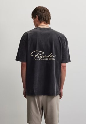 Jeune homme aux cheveux courts porte un T-shirt noir délavé oversize avec le texte « Pegador All Rights Reserved » au dos, short beige.
