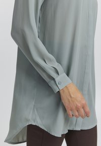 Light blue long-sleeve blouse made of sheer fabric, featuring a buttoned cuff and a slightly curved hem. Smooth texture, minimal design.
