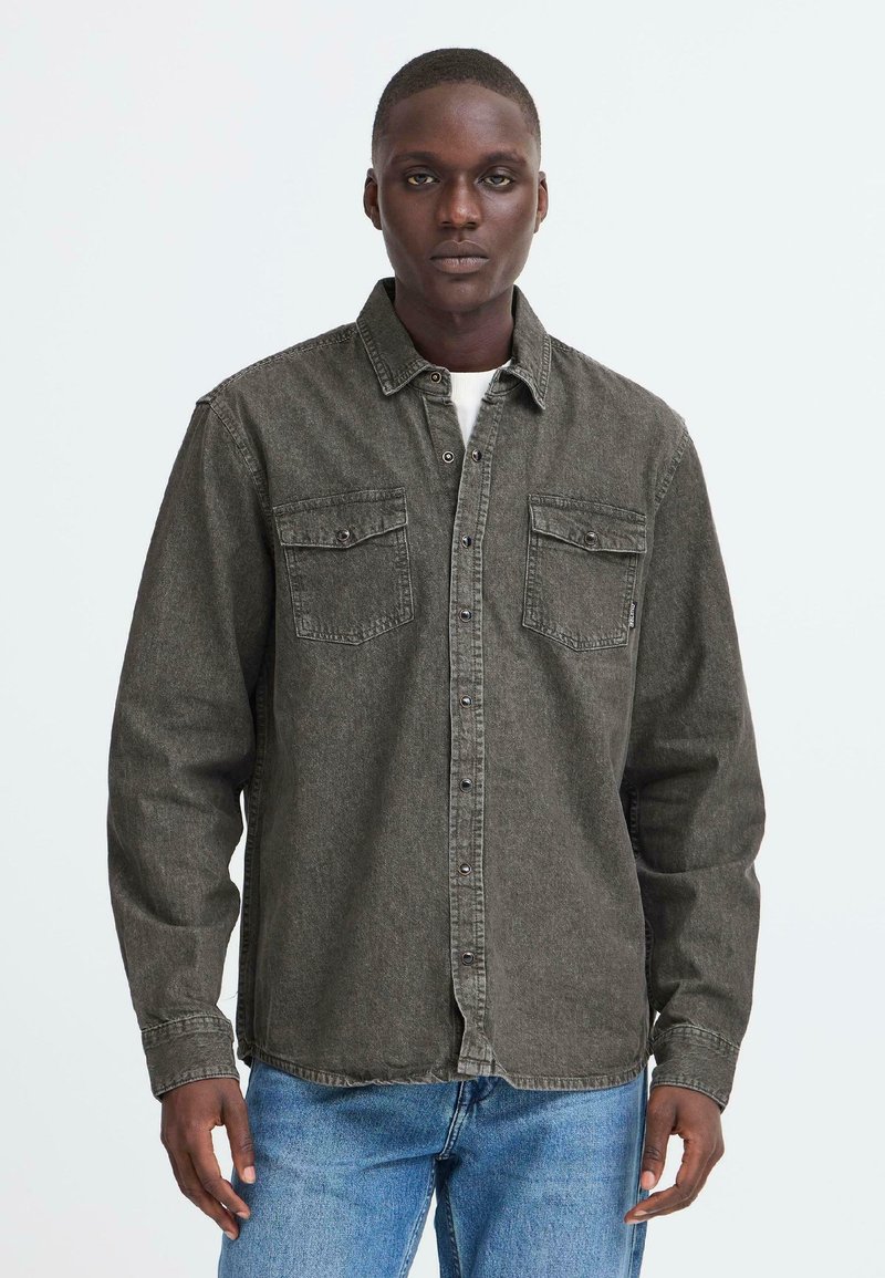 Camicia di denim verde scuro con maniche lunghe, due tasche sul petto e chiusura a bottone, indossata sopra una camicia bianca. Con un taglio finito casuale e rilassato.