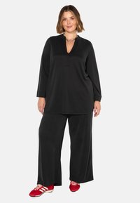 Zwarte, losvallende top met een V-hals en lange mouwen, gecombineerd met wijde zwarte broek. De outfit heeft een lichte stof.