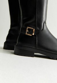 Botas de tobillo de cuero negro con suelas gruesas y detalles de hebilla rectangular dorada en el costado, fotografiadas sobre un fondo blanco.